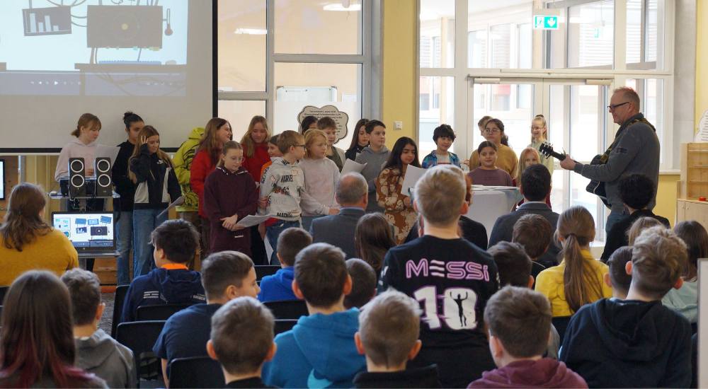 Singende Schüler:innen und ein Mann der Gitarre spielt stehen vor Publikum, das auf Sesseln sitzt.