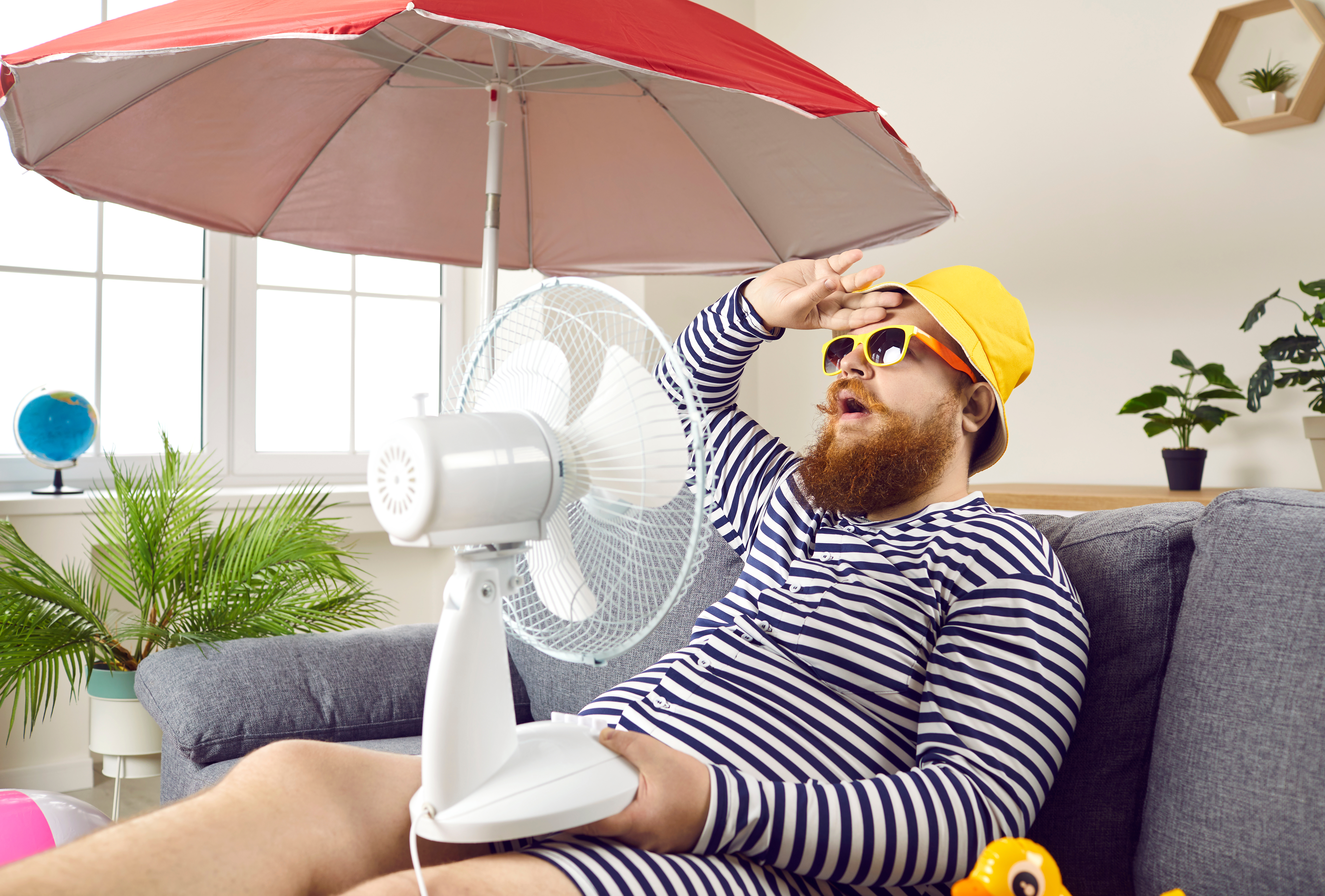 Mann sitzt auf Couch mit gelber sonnenbrillee und Badanzug und hat einen Ventilator vor dem Gesicht.