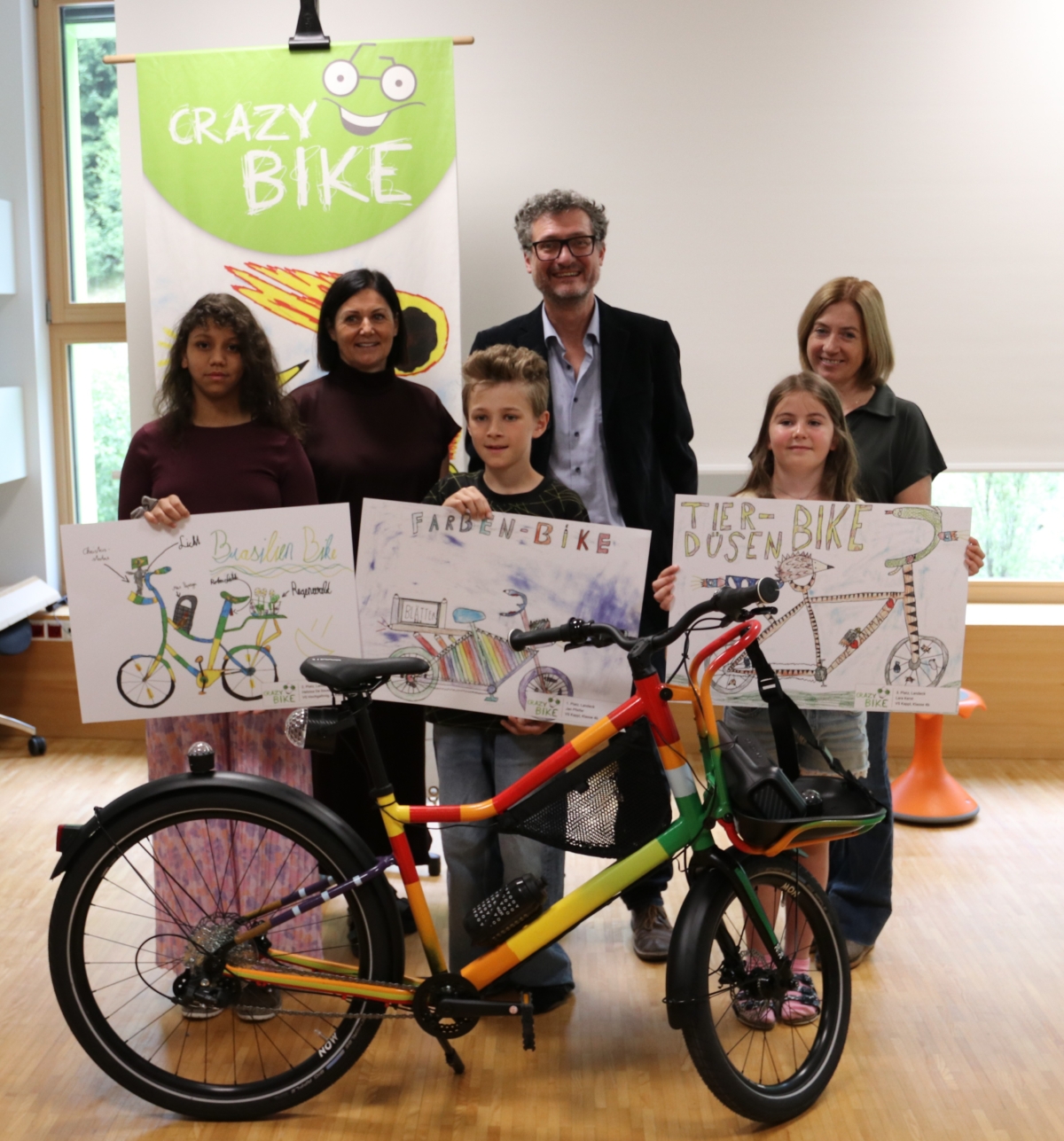 Drei Kinder mit Zeichnungen von Fahrrädern und ein buntes Fahrrad im Vordergrund. Im Hintergrund drei Erwachsene