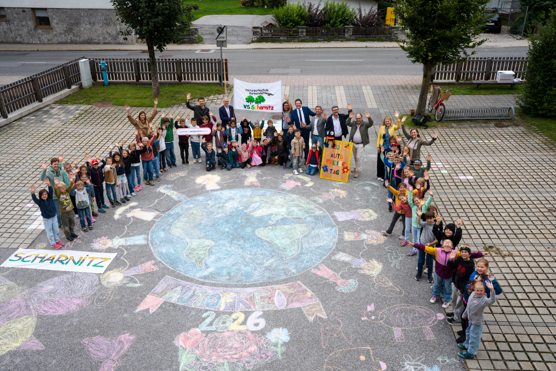 Gruppe von Kindern und Erwachsenen steht im Kreis um eine große bunte Kreidezeichnung der Erde mit dem Schriftzug "EMWTour 2026" auf dem Boden und hält Schilder mit den Aufschriften "VS Scharnitz" und "Alt trifft Neu" hoch